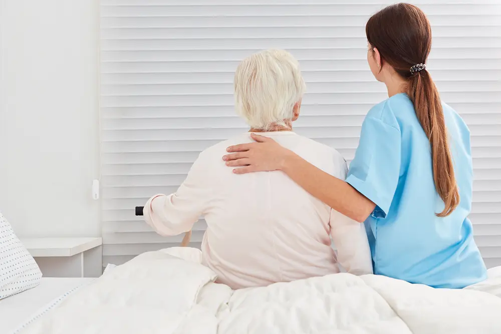 Carer supporting an older person sitting on the edge of a bed, illustrating BedSense real-time bed occupancy monitoring and resident awareness.